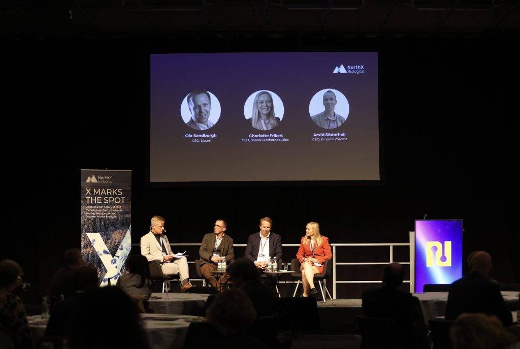 Four people seated on a stage during a panel discussion. One man is moderating the conversation, interviewing two men and one woman under stage lighting.